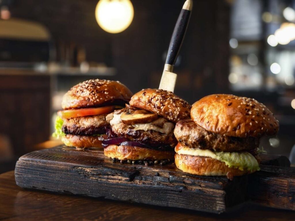 3 Burgers on a wood plank with a knife sticking up out of the middle one for article Best Burger Joints in Austin: 10 Newcomer Must-Tries for those moving to Austin.