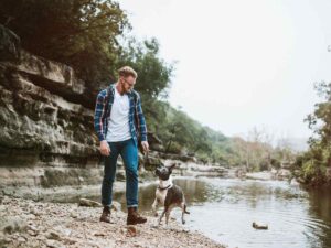 Man hiking in jeans, boots, white t-shirt and flannel long sleeve shirt with his do on the edge of Austin, Texas river for article 12 Great Winter Activities in Austin for newcomers moving to Austin.