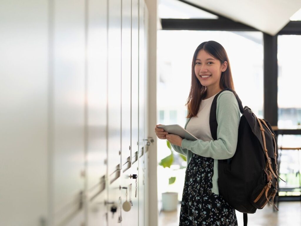A female high school student standing in front of her school locker holding her books with back pack on - for article Private High Schools in Austin for newcomers moving to Austin.