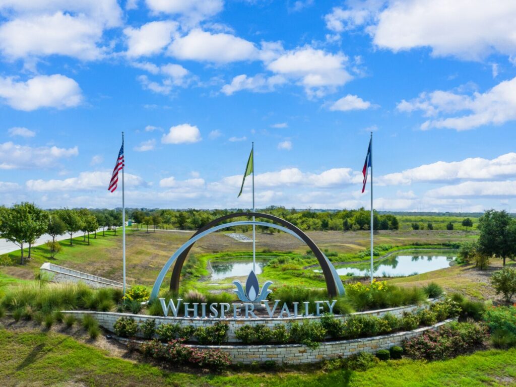 Welcome entrance sign to Whisper Valley community in East Austin, with logo, 3 flags, green grass and blue sky