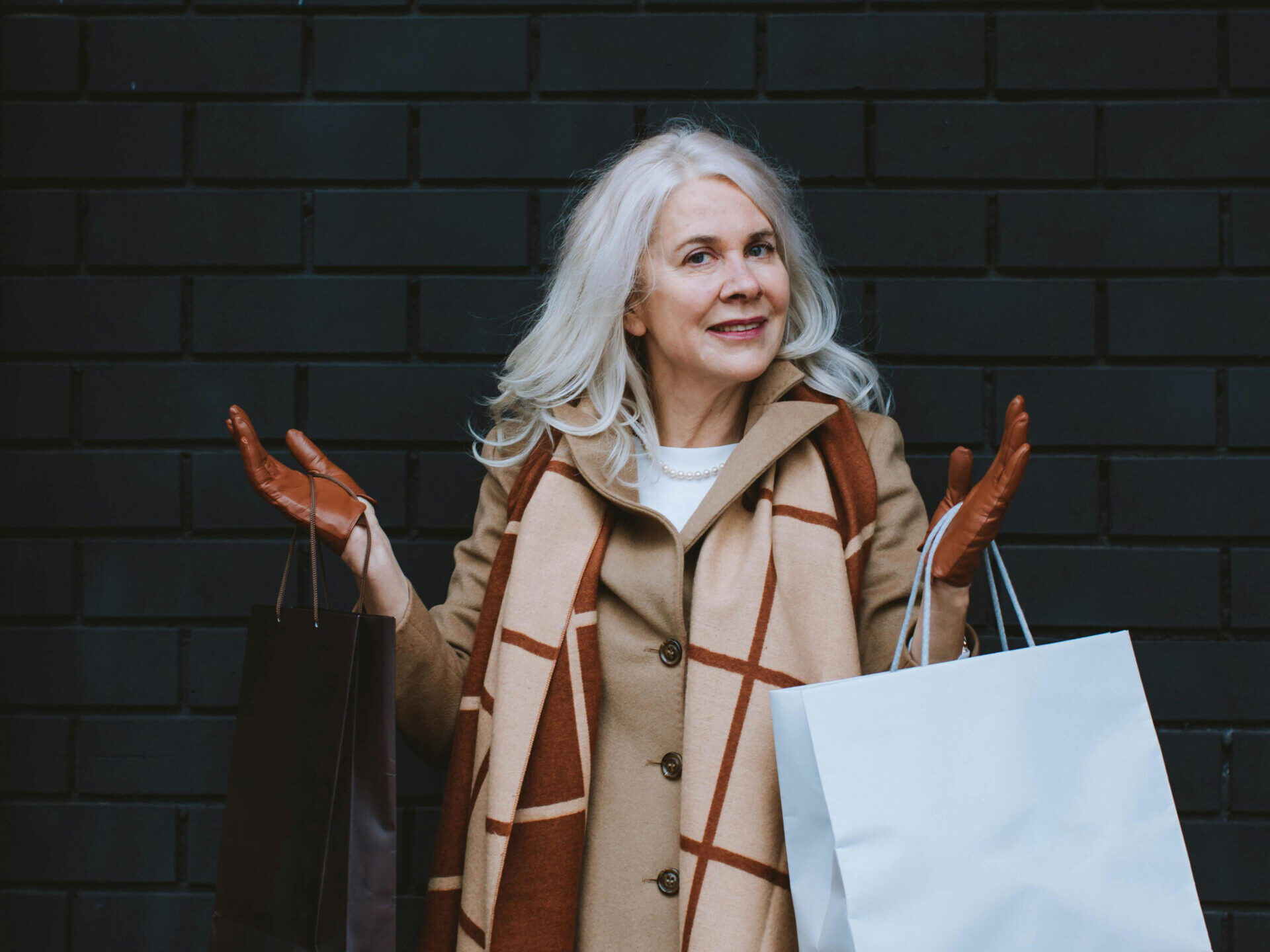 woman with white hair, wearing gloves and winter coat holding shopping bag in front of gray brick wall