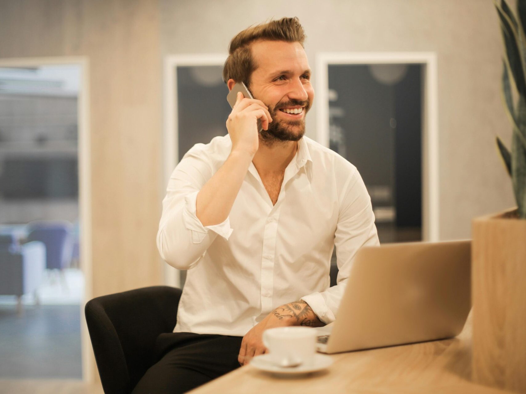 man with beard sitting at desk with laptop, on the phone smiling