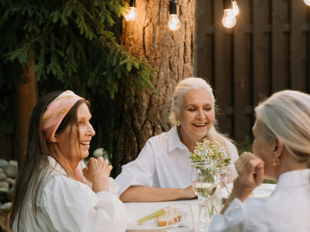 3 women with white hair at an outdoor table, smiling, having wine under string lights