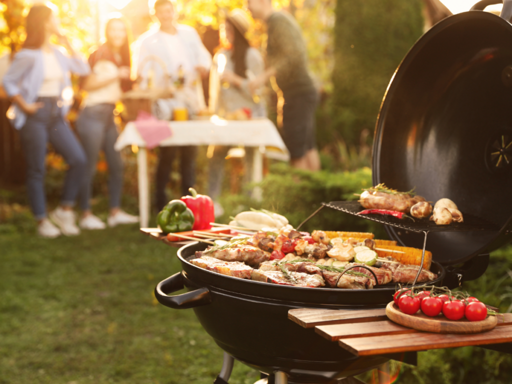 Grill with food on it and people gathered in background.