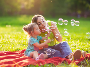 Mom and child on picnic blanket in park blowing bubbles.