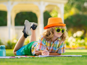 Child laying in green grass wearing summer clothes and sumglasses writing in workbook