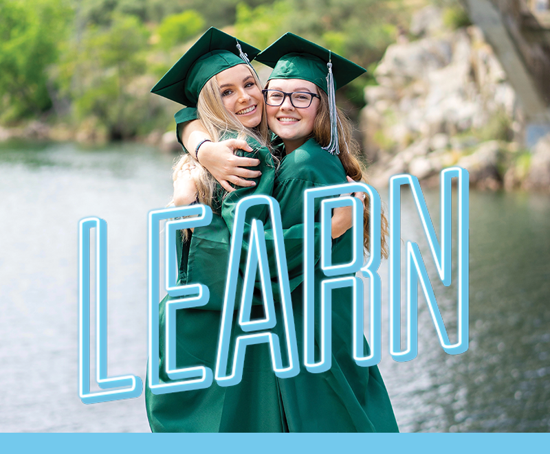 Department Image: LEARN. Young girls with cap and gown hugging near lake.