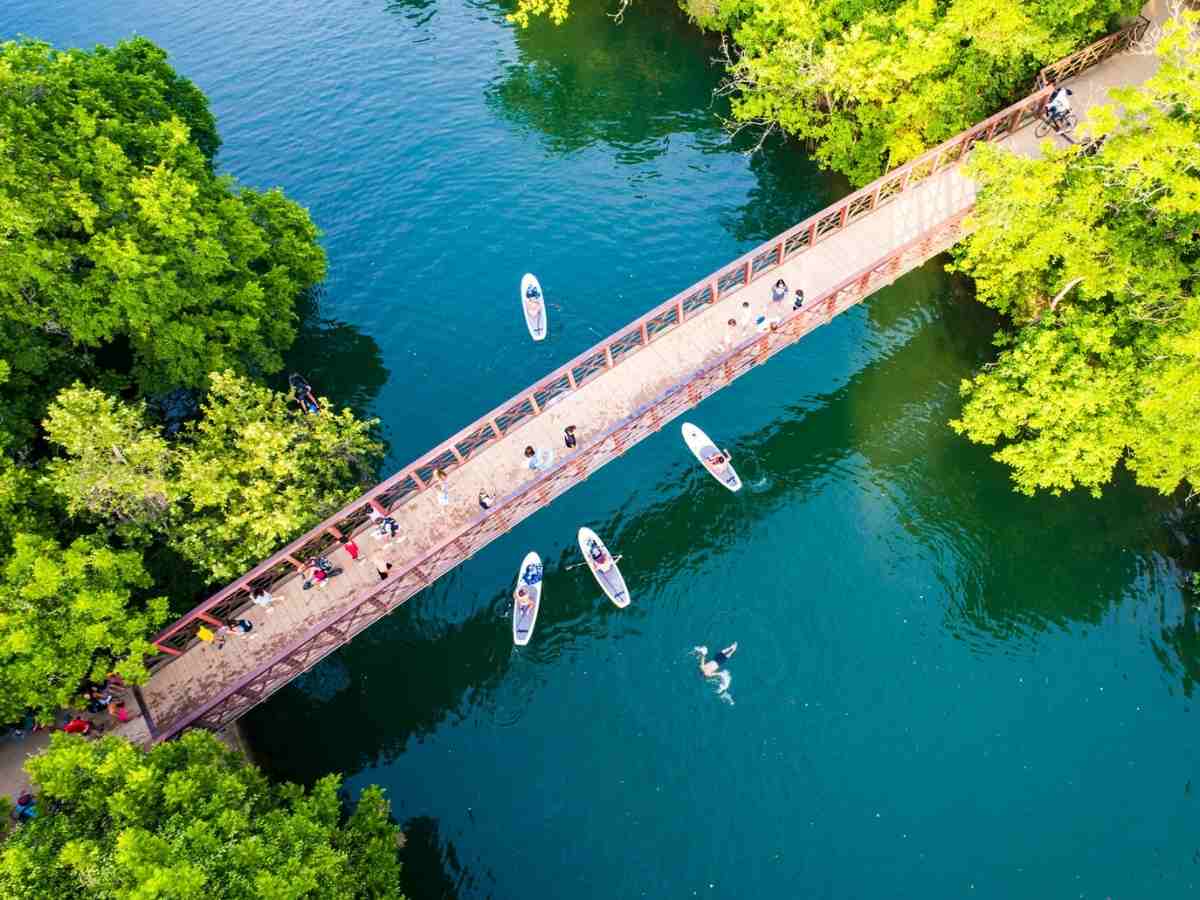 Birds-eye view of river with pedestrian bridge and paddleboards on water. with trees around the edges.