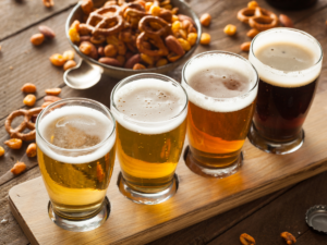 A flight of 4 beers from light to dark on a wood tray with a bowl of bar snacks in background.