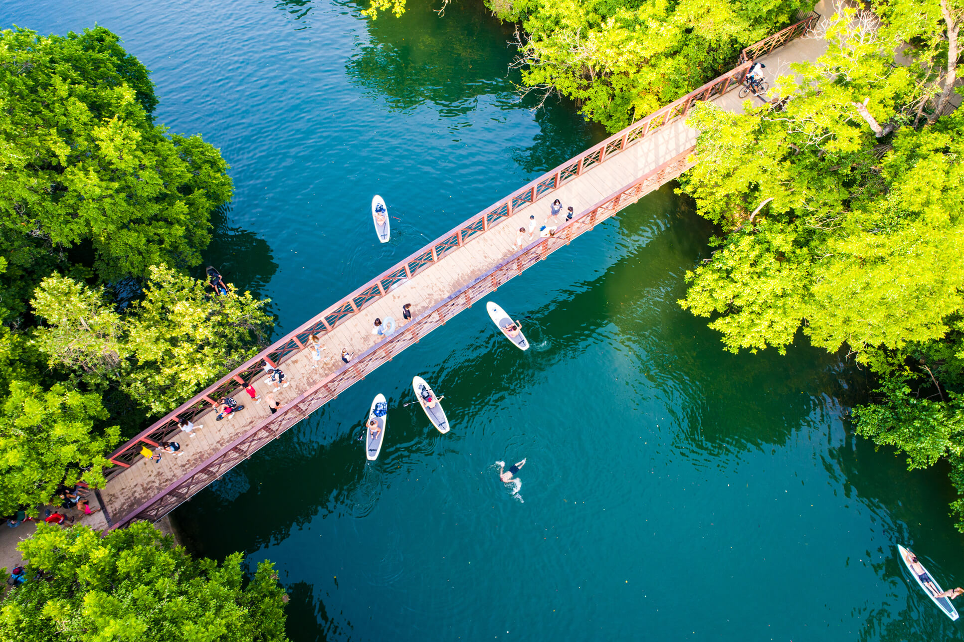 Birds-eye view of river with pedestrian bridge and paddleboards on water. with trees around the edges.