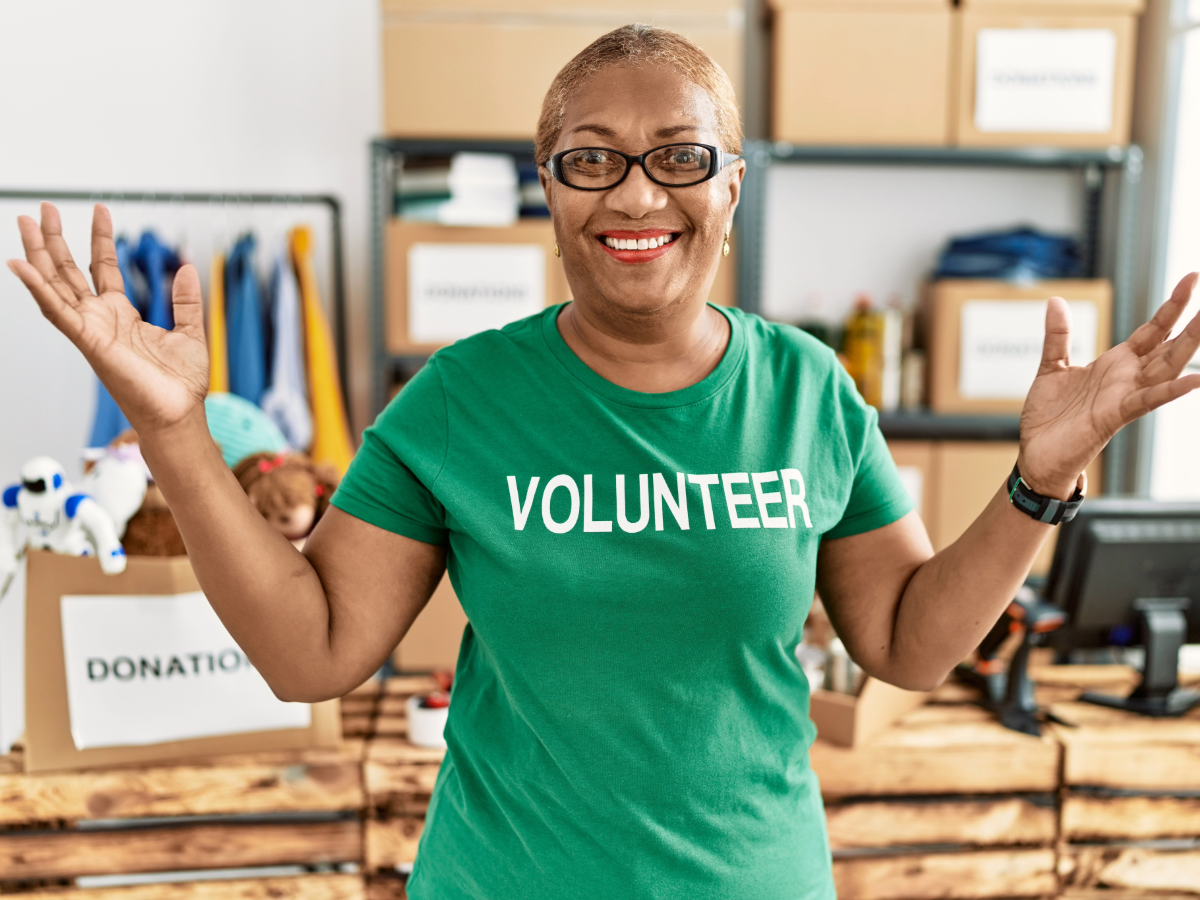 Senior woman in green shirt that says volunteer with donated items behind her.