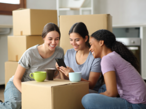 3 female roommates sitting on floor with boxes around them. For Article - Finding Roommates in Austin.