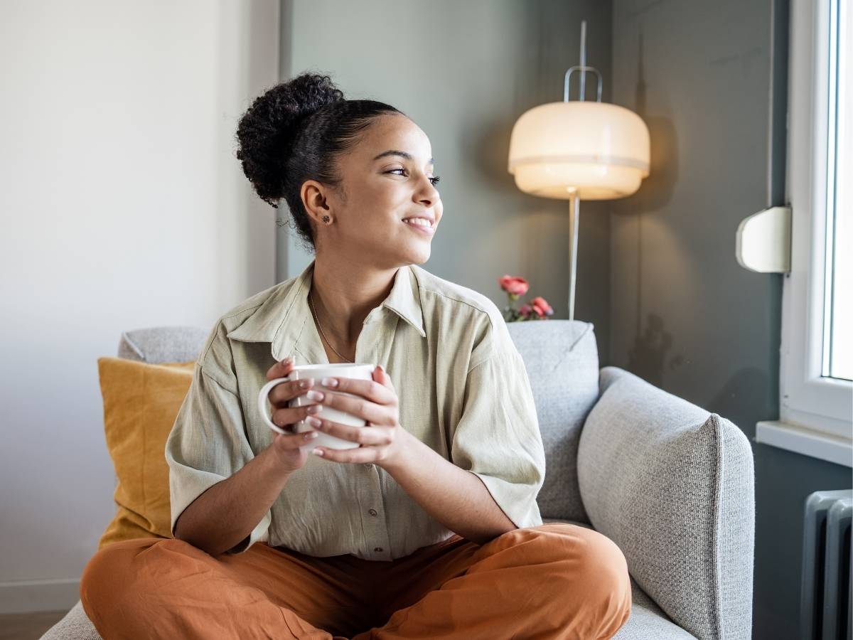 Young African American woman sitting in lotus position on chair with coffee in hand and smiling while looking out window for article Mental Wellness in Austin for newcomers moving to Austin.