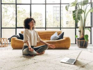 Woman sitting in lotus position on floor of room with couch and laptop open in front of her for article 10 Great Stress Management Programs in Austin for newcomers moving to Austin.