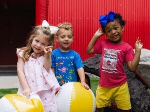 3 preschool kids posing for camera with yellow and white beach balls and a red wall behind them for article Little Thinkers Preschool for newcomers moving to Austin.