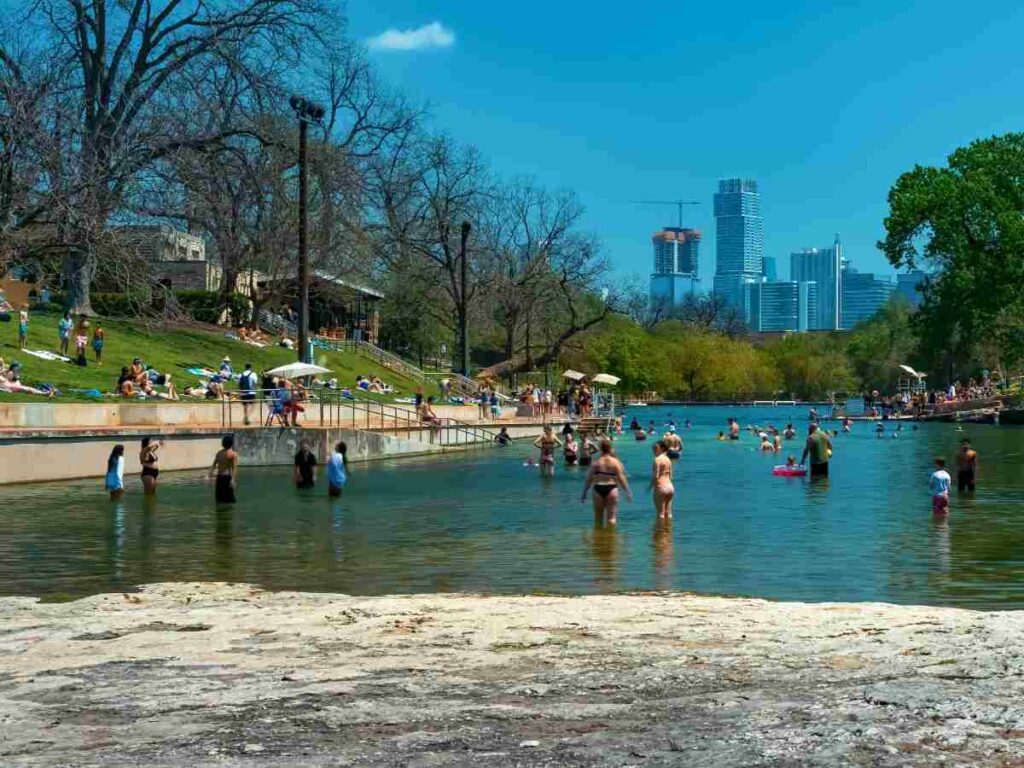 People in the water with Austin skyline in the background for article Everything You Need to Know About Barton Springs Pool for newcomers moving to Austin.